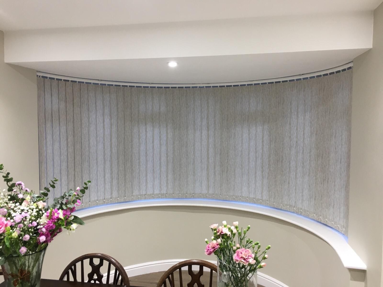 Curved Vertical Blinds in Vivi Stone – Made to Measure in Essex Made-to-measure curved vertical blinds in Vivi Stone fabric, installed in a bay window dining area of an Essex home, offering smooth operation and soft light control.