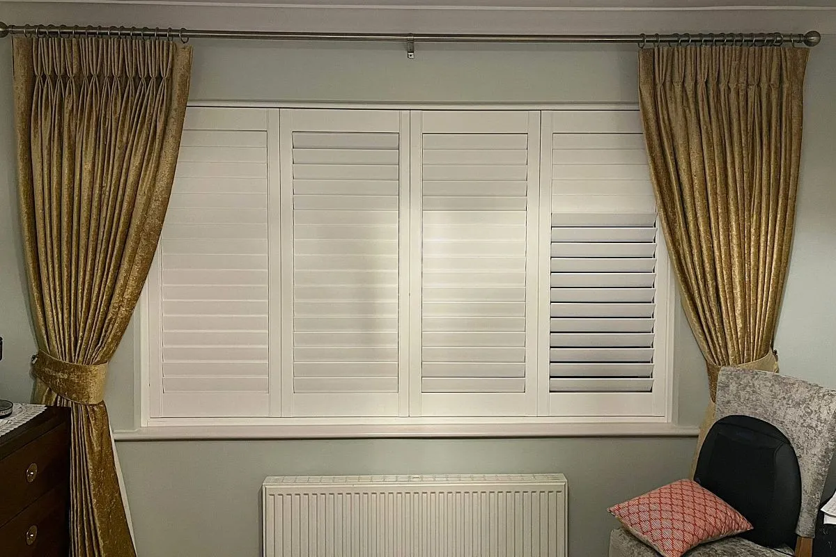 Gold textured pencil pleat curtains on an antique brass curtain pole, paired with white plantation shutters in a soft, neutral room.