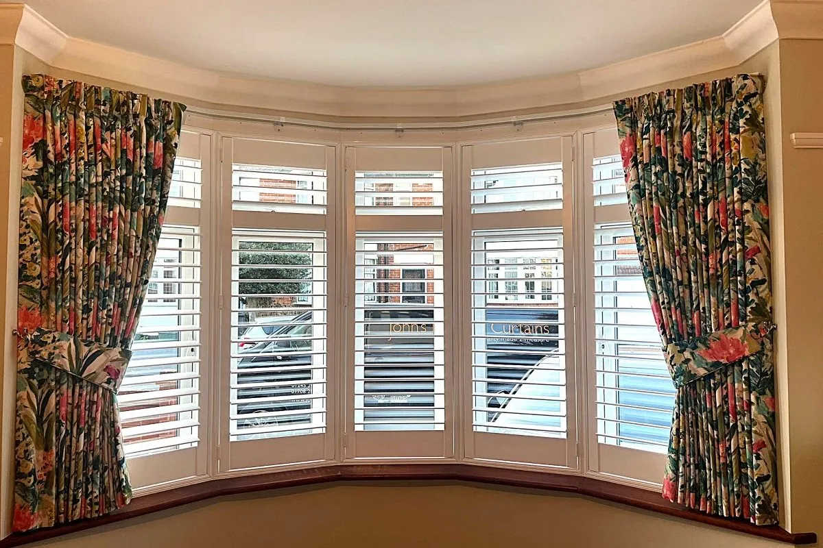 Floral pencil pleat curtains on a bay window with white plantation shutters, creating a bright and elegant living space.