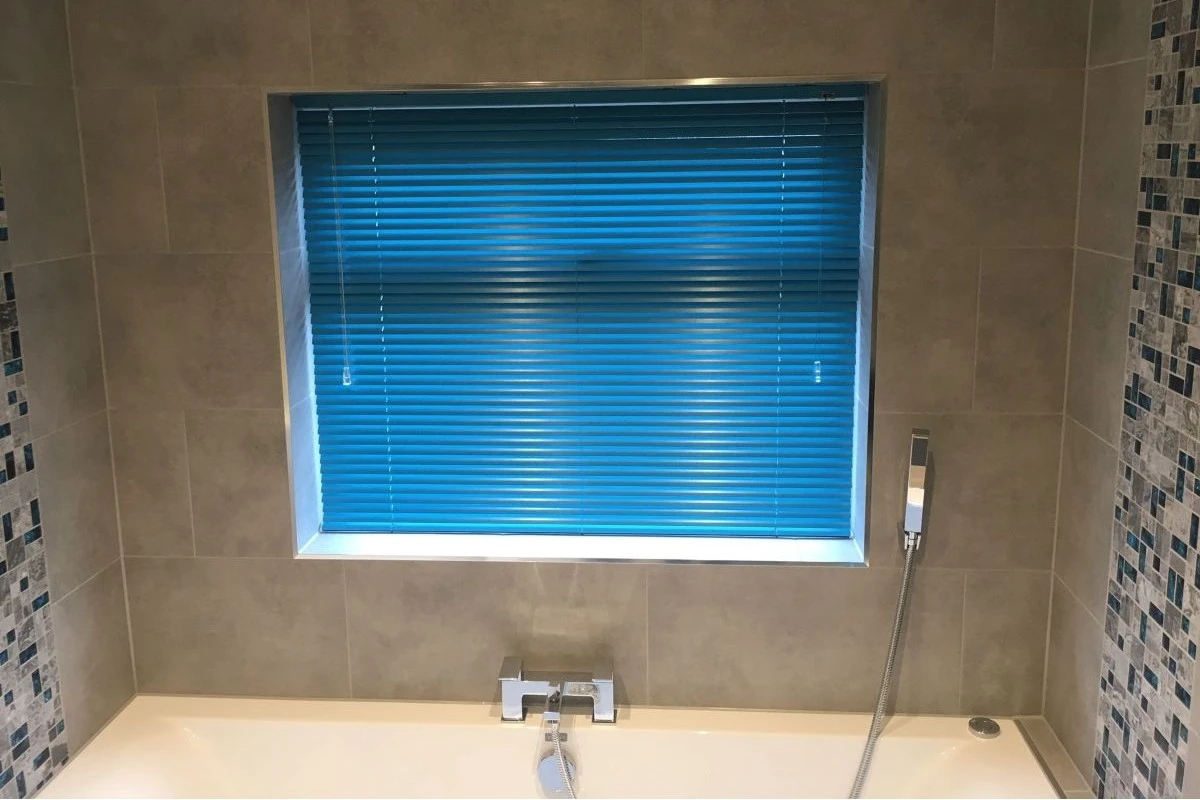 Blue aluminium Venetian blinds fitted above a bath in a tiled bathroom, providing waterproof privacy and adjustable light control.