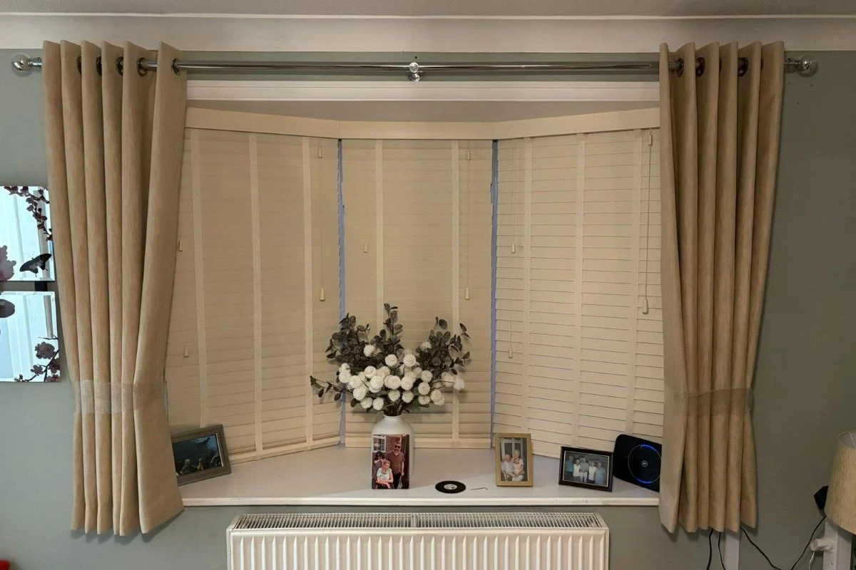 Beige eyelet curtains fitted on a bay window with plantation shutters, installed by John’s Blinds & Curtains.