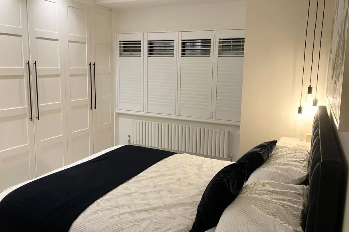 Full-Height Plantation Shutters in a Modern Bedroom Full-height white plantation shutters fitted across a bedroom window, shown in a modern room with built-in wardrobes, warm lighting and a neatly dressed bed.