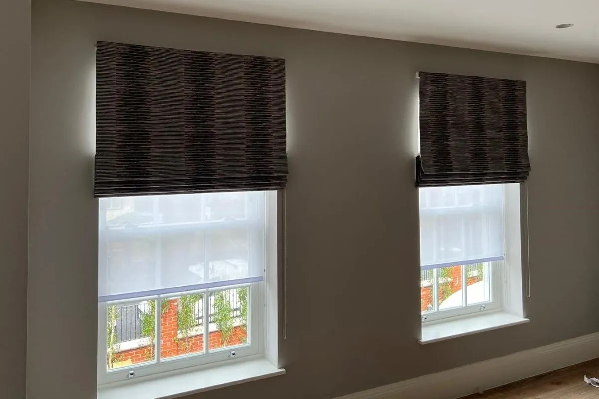 Two windows fitted with patterned Roman blinds layered over sheer white voile roller blinds, providing privacy and light control.