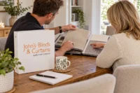 Two people looking through fabric samples at home, with a John’s Blinds & Curtains folder on the table.